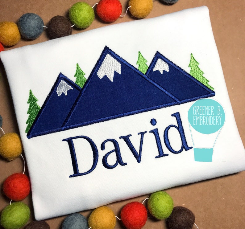May include: A white t-shirt featuring a blue mountain range design with green trees. The name "David" is embroidered beneath the mountains.
