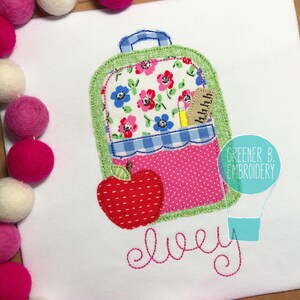 May include: A green and pink backpack with a red apple and a pencil in the pocket. The backpack has a floral pattern and the name "elvey" is embroidered below it.