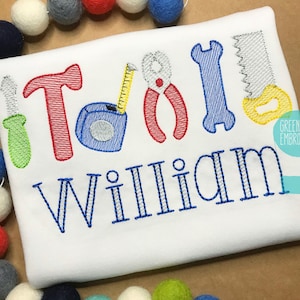 May include: White t-shirt embroidered with colorful tool designs spelling "TOOL" above the name "WILLIAM" in blue. The tools include a screwdriver, hammer, tape measure, pliers, wrench, and saw. The shirt is surrounded by colorful felt balls.