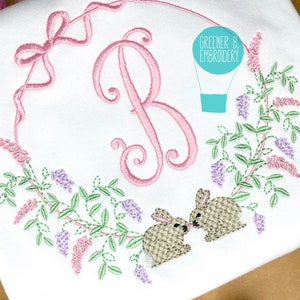 May include: An embroidered design featuring a pink letter B surrounded by a wreath of green leaves and purple and pink flowers. Two brown bunnies are kissing on the bottom of the wreath. The text "Greener B. Embroidery" is in a blue circle above the wreath.
