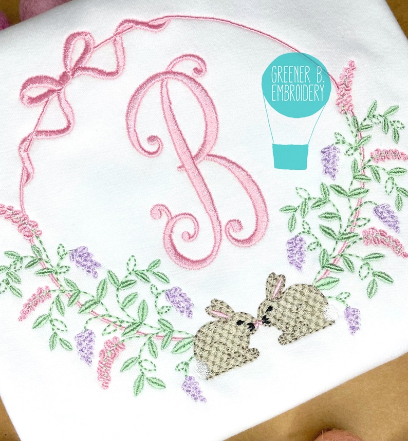 May include: An embroidered design featuring a pink letter B surrounded by a wreath of green leaves and purple and pink flowers. Two brown bunnies are kissing on the bottom of the wreath. The text "Greener B. Embroidery" is in a blue circle above the wreath.