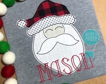 May include: A gray t-shirt featuring a Santa Claus face design. The Santa hat is red and black buffalo plaid, with a white polka dot trim. The name "MASON" is embroidered in red below the face. A garland of red, white, and green felt balls is in the background.