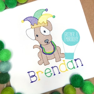 May include: Embroidered design of a brown dog wearing a purple, green, and yellow jester hat and a green, purple, and gold beaded necklace. The dog is sitting and holding a light blue hot air balloon. The name "Brendan" is embroidered below the dog in green, yellow, and purple thread. The text "Greener B. Embroidery" is embroidered on the balloon.