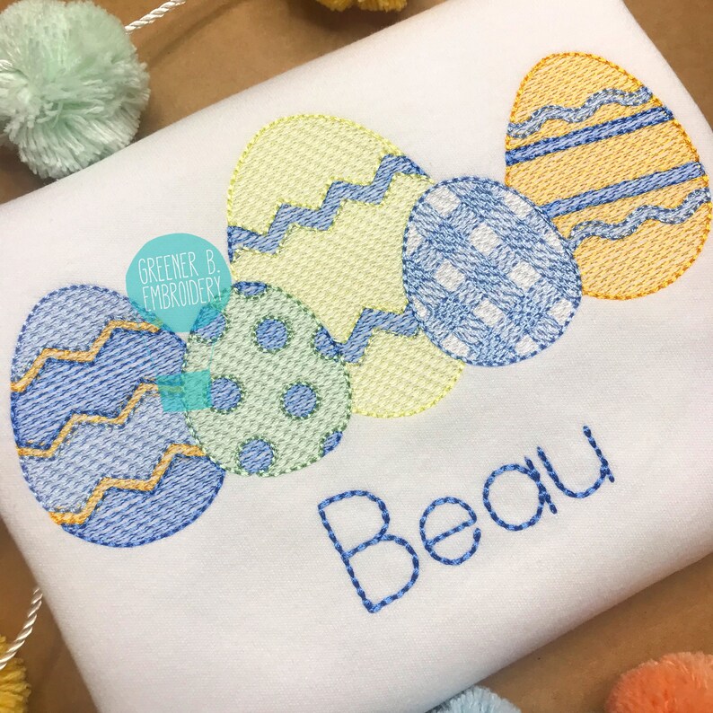 以下が含まれることがあります： 白い刺繍の入ったシャツに、カラフルなイースターエッグが5つと、青い糸で「Beau」という名前が刺繍されています。