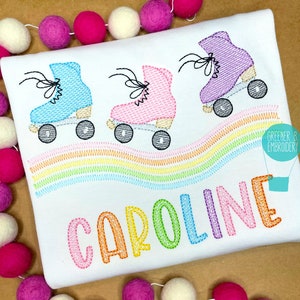 May include: A white t-shirt with a rainbow design and three roller skates in blue, pink, and purple. The name "CAROLINE" is embroidered in rainbow colors below the roller skates.