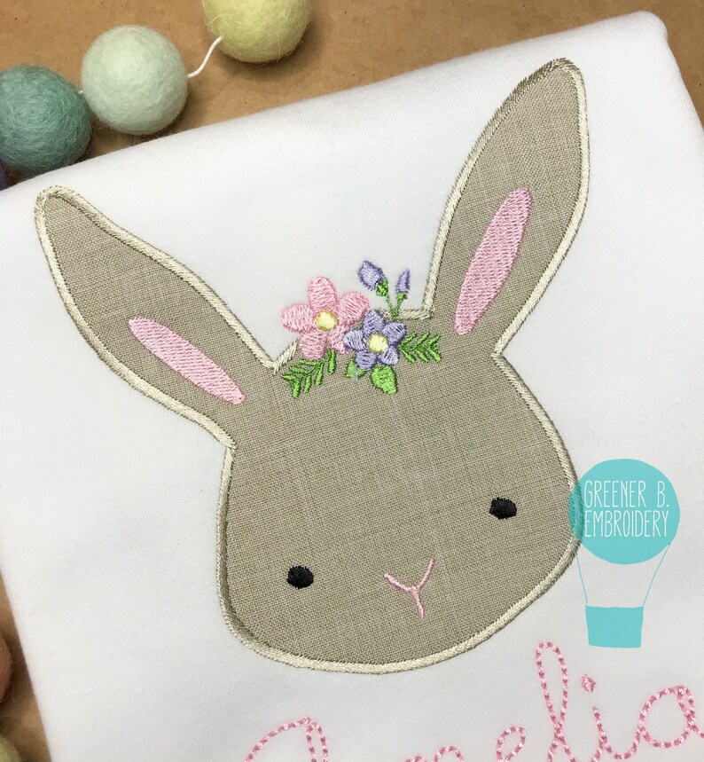 May include: Embroidered bunny head with a floral crown in shades of pink, purple, and green. The bunny has a brown face with black eyes and a pink nose. The text "Greener B. Embroidery" is visible in the bottom right corner.