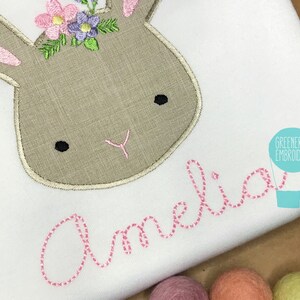 May include: A white onesie with a brown embroidered bunny wearing a pink flower crown. The name "Amelia" is embroidered in pink below the bunny.