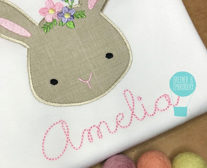 May include: A white onesie with a brown embroidered bunny wearing a pink flower crown. The name "Amelia" is embroidered in pink below the bunny.