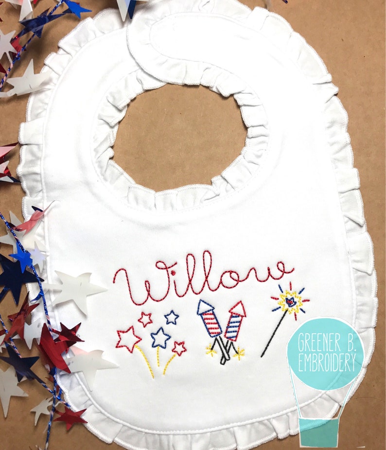 May include: White baby bib with a red, white, and blue embroidered design of fireworks and stars. The name "Willow" is embroidered in red above the design. The bib has a ruffled edge.