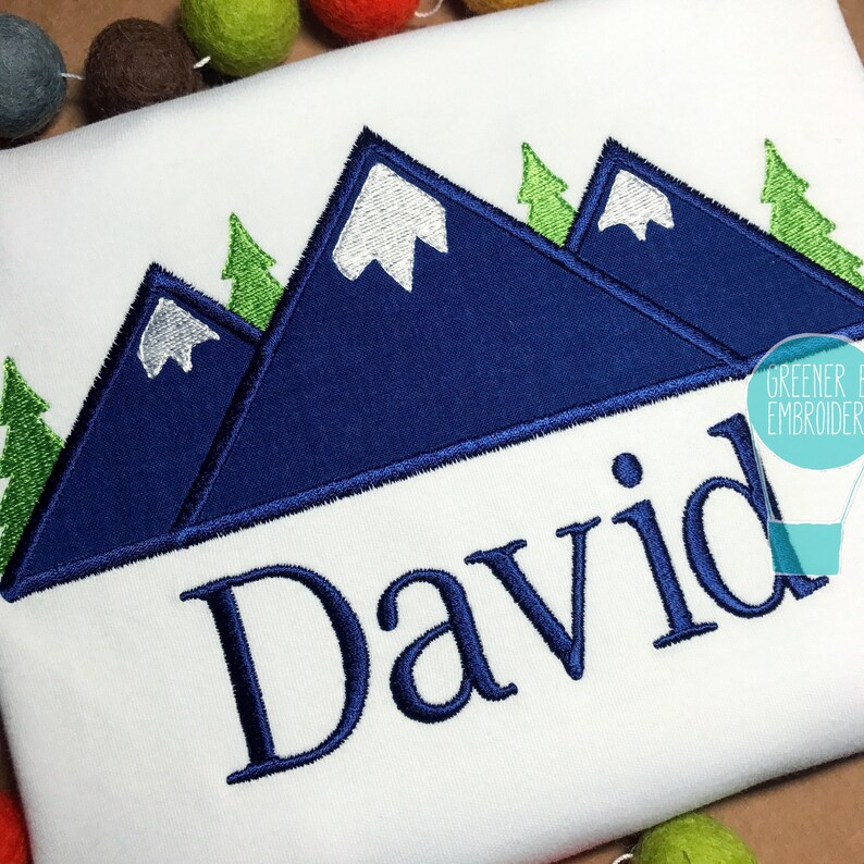 May include: A white t-shirt with a blue mountain range design and the name "David" embroidered in blue below. The mountains have white snow peaks and green trees.