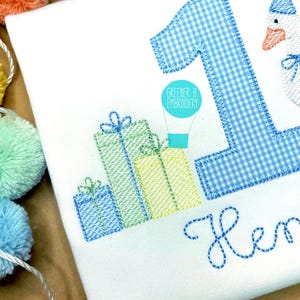 May include: A white onesie with blue and green embroidered presents, a blue gingham number one, a hot air balloon, and the name "Henry".