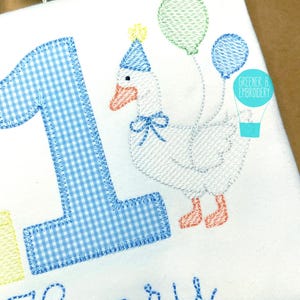May include: A white embroidered goose wearing a blue party hat and holding balloons. The goose is next to a blue and white gingham number one and the name "Henry" is embroidered below.