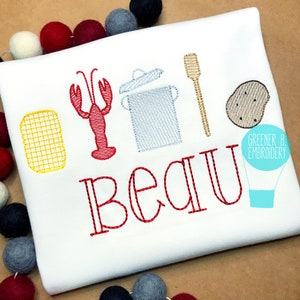 May include: White t-shirt embroidered with a red lobster, cooking pot, spatula, and potato. The name "Beau" is embroidered in red. A yellow grill and a hot air balloon logo are also present.