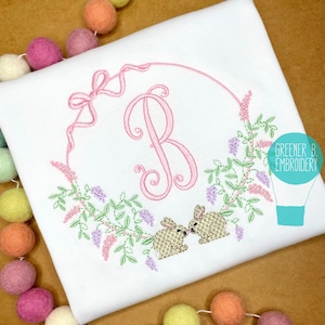 May include: Embroidered design featuring a pink letter B in a wreath of green leaves and purple flowers with two brown bunnies peeking out from behind the wreath. The design is on a white fabric.