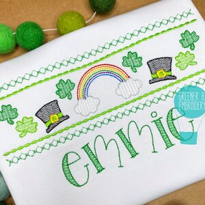 Puede incluir: Una camisa blanca bordada con un tr&eacute;bol verde, un arco&iacute;ris y un sombrero de duende. El nombre "Emme" est&aacute; bordado en verde debajo del dise&ntilde;o.