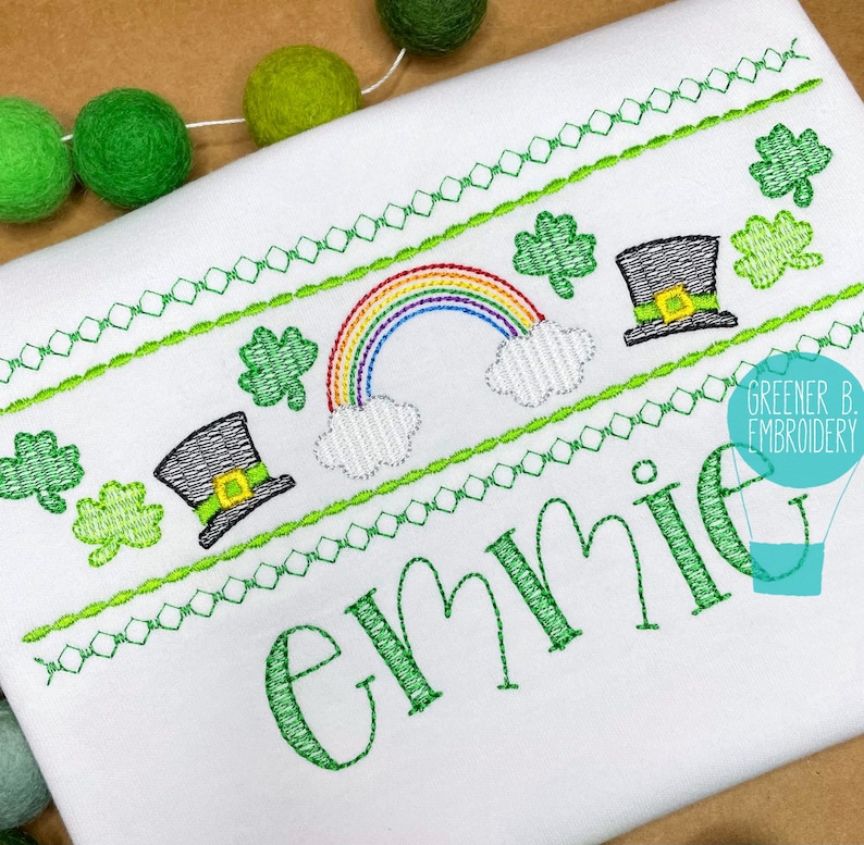 Puede incluir: Una camisa blanca bordada con un tr&eacute;bol verde, un arco&iacute;ris y un sombrero de duende. El nombre "Emme" est&aacute; bordado en verde debajo del dise&ntilde;o.
