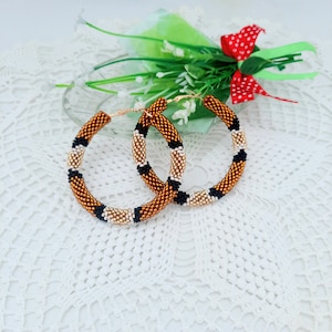 Puede incluir: Un par de pendientes grandes de aro hechos con cuentas marrones, negras y doradas. Los pendientes están sobre una superficie de encaje blanco.