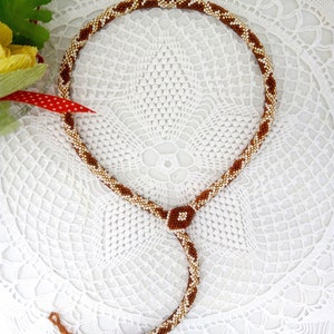 Könnte beinhalten: Eine braune und weiße Perlenkette mit einem quadratischen Perlenakzent. Die Kette liegt auf einer weißen Häkelspitze.