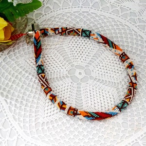 May include: A colorful beaded necklace with a geometric pattern in shades of red, orange, blue, and white. The necklace is made of small beads and has a silver clasp.