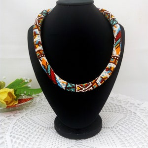 May include: A colorful beaded necklace with a geometric pattern in shades of white, brown, orange, blue, and turquoise. The necklace is displayed on a black mannequin.