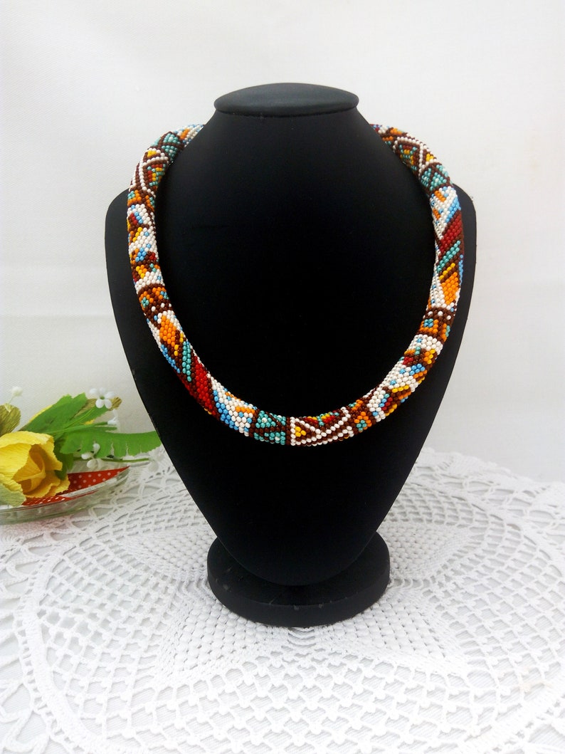 May include: A colorful beaded necklace with a geometric pattern in shades of white, brown, orange, blue, and turquoise. The necklace is displayed on a black mannequin.