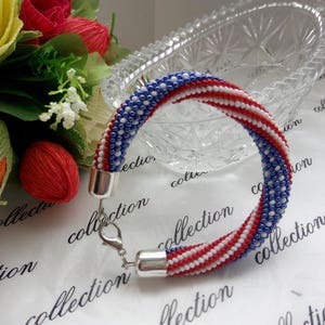 Peut inclure: Un bracelet en perles avec des rayures rouges, blanches et bleues, rappelant le drapeau fran&ccedil;ais. Le bracelet est fait de petites perles et a un fermoir argent&eacute;.