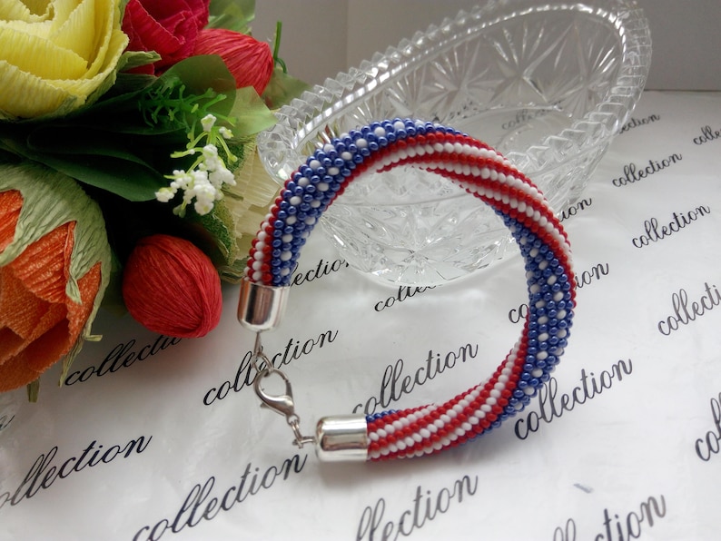 Peut inclure: Un bracelet en perles avec des rayures rouges, blanches et bleues, rappelant le drapeau fran&ccedil;ais. Le bracelet est fait de petites perles et a un fermoir argent&eacute;.
