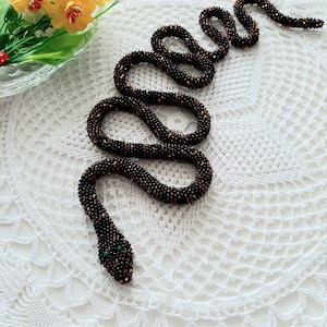 May include: A beaded snake made of black and brown beads with green eyes. The snake is coiled on a white crocheted surface.