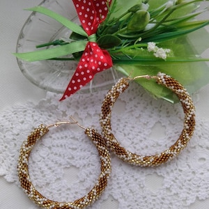 Puede incluir: Un par de grandes pendientes de aro de cuentas doradas y blancas. Los pendientes están hechos con pequeñas cuentas redondas que se tejen juntas en un patrón. Los pendientes están colgando sobre un fondo de encaje blanco.