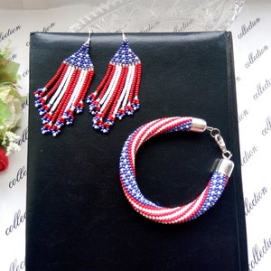 Peut inclure: Un ensemble de bijoux en perles de couleur rouge, blanc et bleu. L'ensemble comprend un bracelet et une paire de boucles d'oreilles pendantes. Les bijoux sont faits de petites perles et ont un th&egrave;me patriotique.