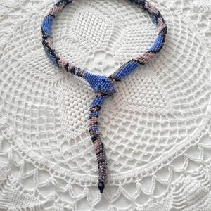 Puede incluir: Un collar de serpiente de cuentas azules, rosas y negras. El collar está enrollado en un círculo y luego cuelga en forma de serpiente.