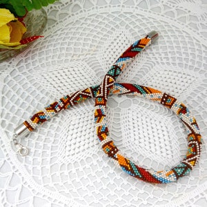 May include: A colorful beaded necklace with a geometric pattern in shades of brown, orange, red, blue, and white. The necklace is made of small beads and has a silver clasp.