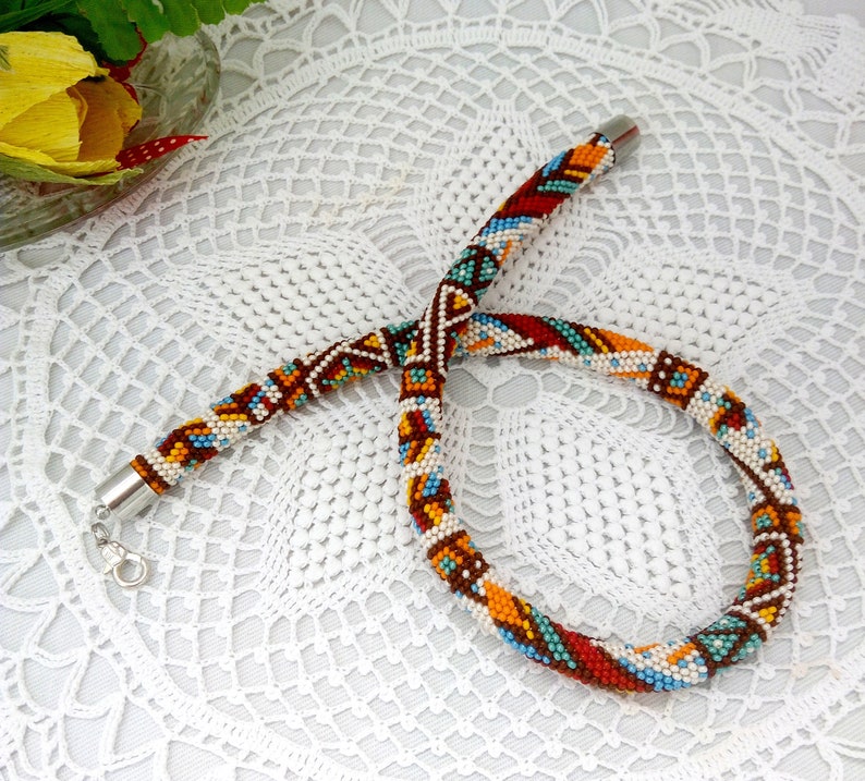 May include: A colorful beaded necklace with a geometric pattern in shades of brown, orange, red, blue, and white. The necklace is made of small beads and has a silver clasp.