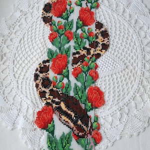 May include: A beaded necklace featuring a snake and red flowers design. The necklace is made with green and white beads and has a fringe of green beads.