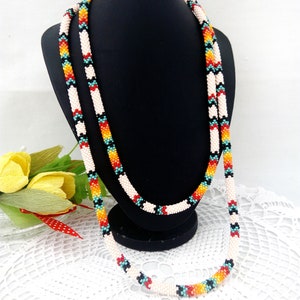 Puede incluir: Un collar largo de cuentas con una base blanca y patrones geométricos coloridos en rojo, naranja, amarillo, turquesa y negro.