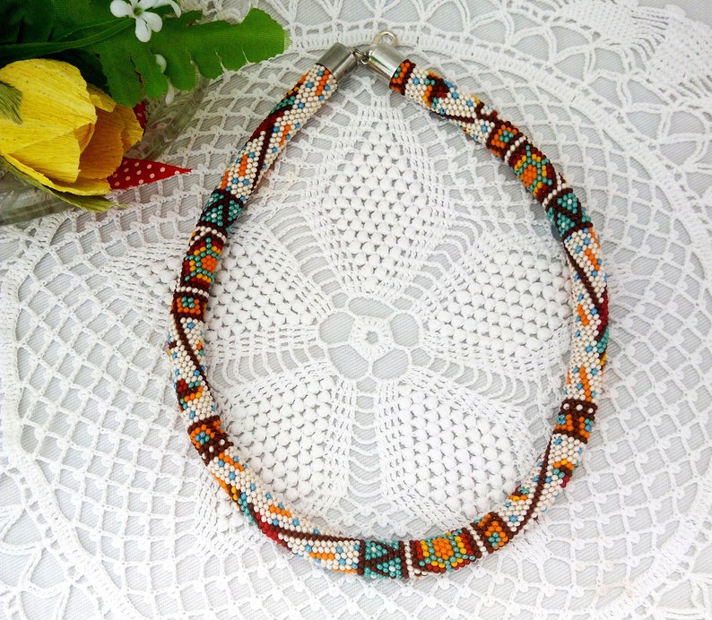 May include: A colorful beaded necklace with a geometric pattern in shades of brown, orange, white, and blue. The necklace is made of small beads and has a silver clasp.