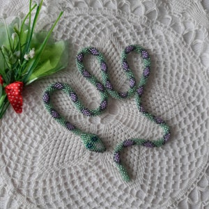 Puede incluir: Un collar de serpiente de cuentas verdes y moradas con un ojo verde y azul. La serpiente está enroscada sobre una superficie blanca de ganchillo.