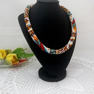May include: A colorful beaded necklace with geometric patterns in shades of brown, orange, white, and blue. The necklace is displayed on a black mannequin.