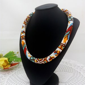 May include: A colorful beaded necklace with a geometric pattern in shades of brown, orange, red, white, and blue. The necklace is displayed on a black mannequin.