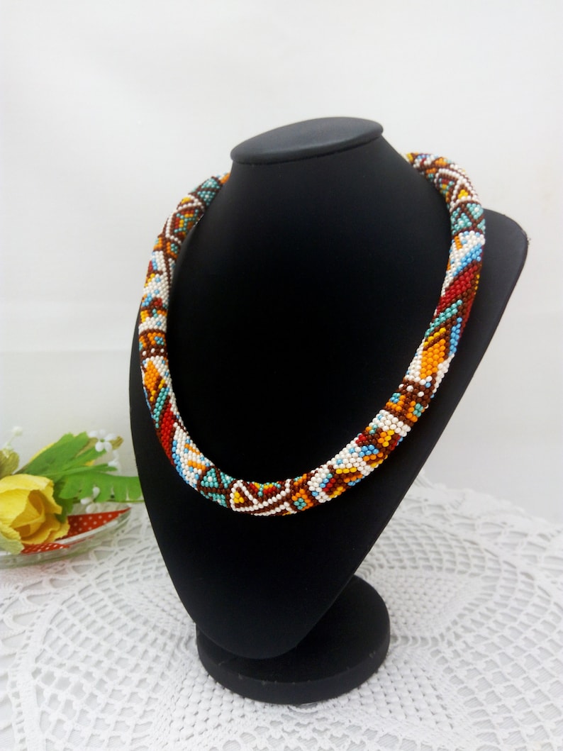 May include: A colorful beaded necklace with a geometric pattern in shades of brown, orange, red, white, and blue. The necklace is displayed on a black mannequin.