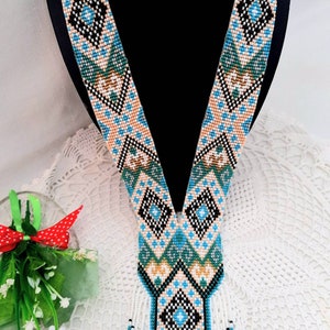 May include: A beaded necklace with a geometric pattern in shades of blue, green, black, and white. The necklace has a V-shaped design and a fringe at the bottom.