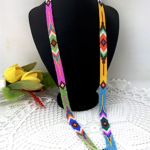 May include: A long beaded necklace with a geometric pattern in shades of pink, green, blue, orange, and yellow. The necklace is displayed on a black mannequin.