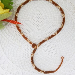 Könnte beinhalten: Eine braune und goldfarbene Perlenkette mit einem schlangenartigen Design. Die Halskette besteht aus kleinen, runden Perlen und hat einen zentralen Knoten. Die Halskette wird auf einer weißen, spitzenartigen Oberfläche präsentiert.