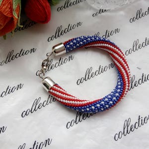 Peut inclure: Un bracelet en perles avec des perles rouges, blanches et bleues dispos&eacute;es en un motif ressemblant au drapeau am&eacute;ricain. Le bracelet est fait de petites perles rondes et a un fermoir argent&eacute;.
