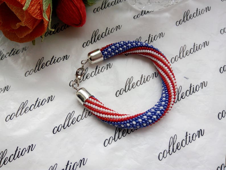 Peut inclure: Un bracelet en perles avec des perles rouges, blanches et bleues dispos&eacute;es en un motif ressemblant au drapeau am&eacute;ricain. Le bracelet est fait de petites perles rondes et a un fermoir argent&eacute;.