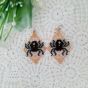 Puede incluir: Un par de pendientes de cuentas con un diseño de araña negra sobre un fondo blanco y dorado.