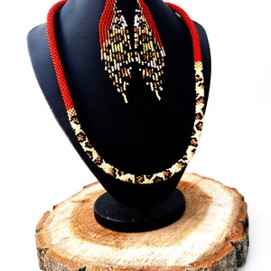 May include: A red and brown beaded necklace and matching earrings. The necklace is on a black display stand and the earrings are hanging from the stand. The necklace and earrings are made of small beads in a geometric pattern.