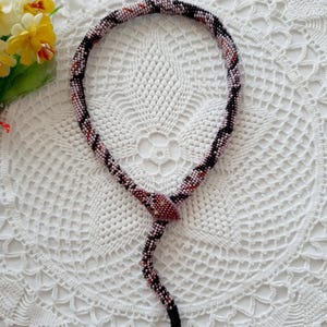 Op de afbeelding: Een kralen slangenketting met een roze, zwart en oranje kleurenschema. De ketting ligt op een wit gehaakt doily.