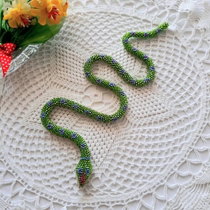 May include: A green beaded snake with blue and red accents. The snake is curled up on a white crocheted doily.