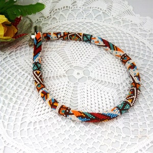 May include: A colorful beaded necklace with a geometric pattern in shades of red, orange, blue, white, and brown. The necklace is made of small beads and has a clasp closure.